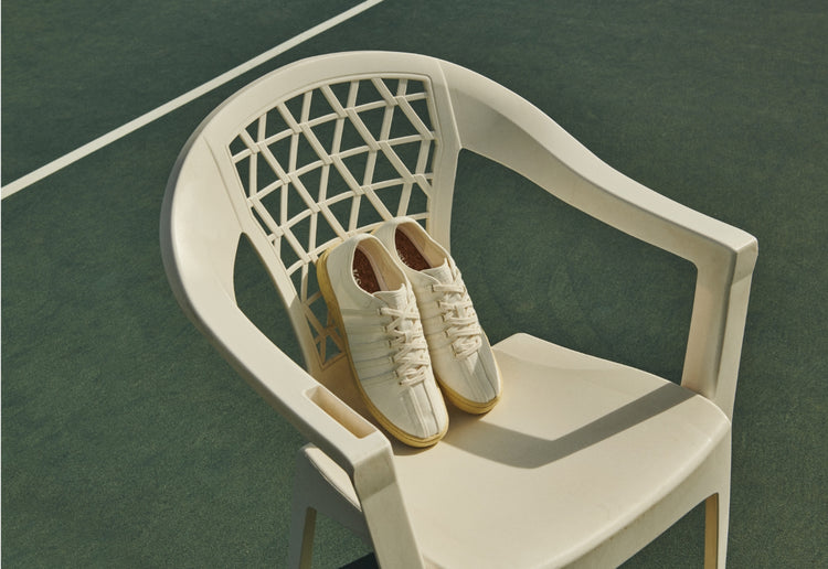 White sporty K-Swiss Corridor collection shoes on a white chair, set on a green tennis court.
