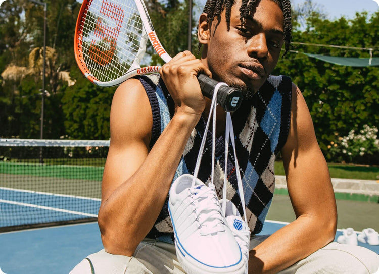 Image of a man holding a tennis racket with a pair of white K-Swiss shoes tied around the handle