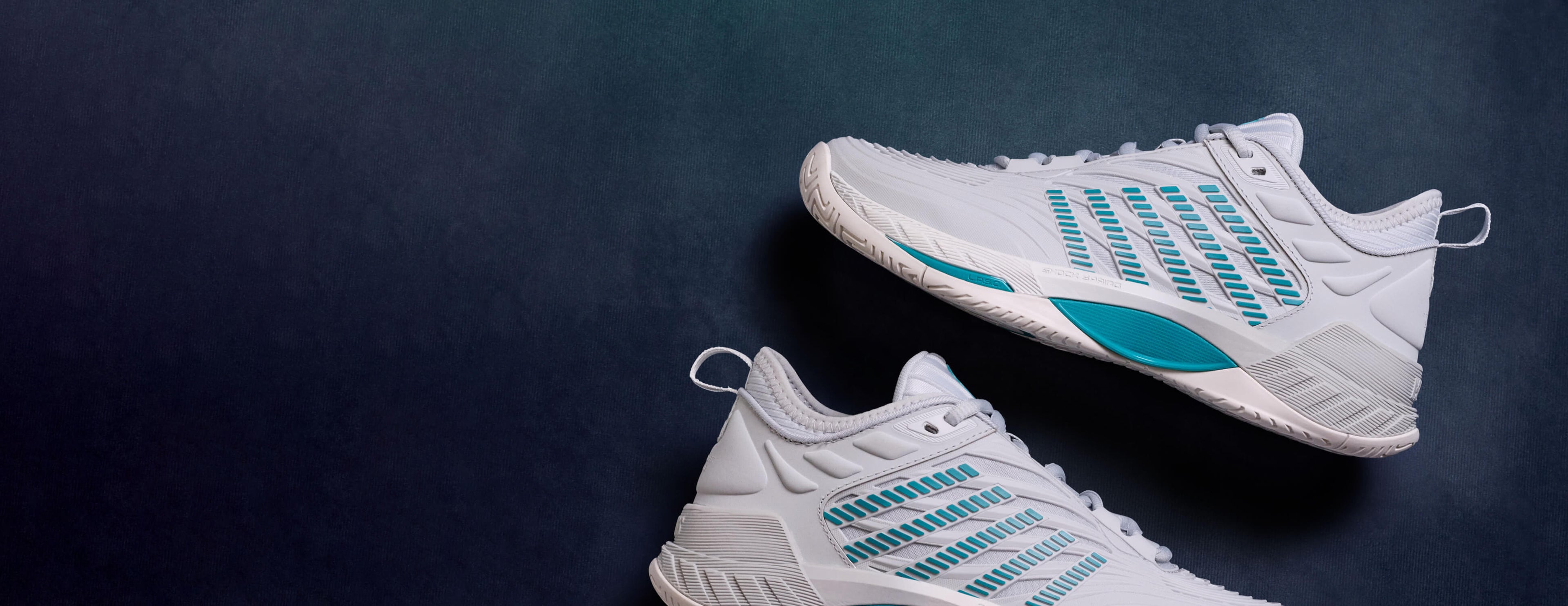 A pair of white athletic sneakers with teal accents, displayed diagonally on a dark textured background.