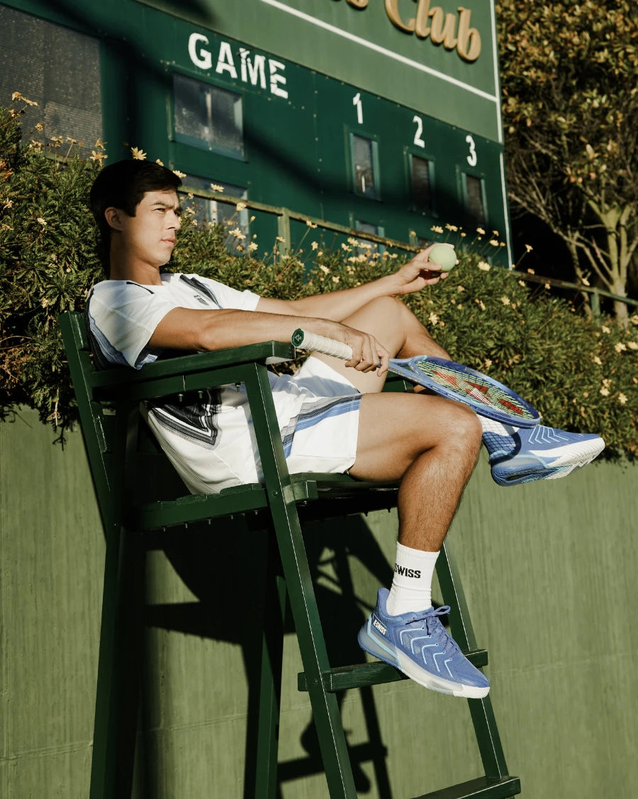 Person sitting on a high chair with sports equipment, holding a tennis ball, in front of a scoreboard.
