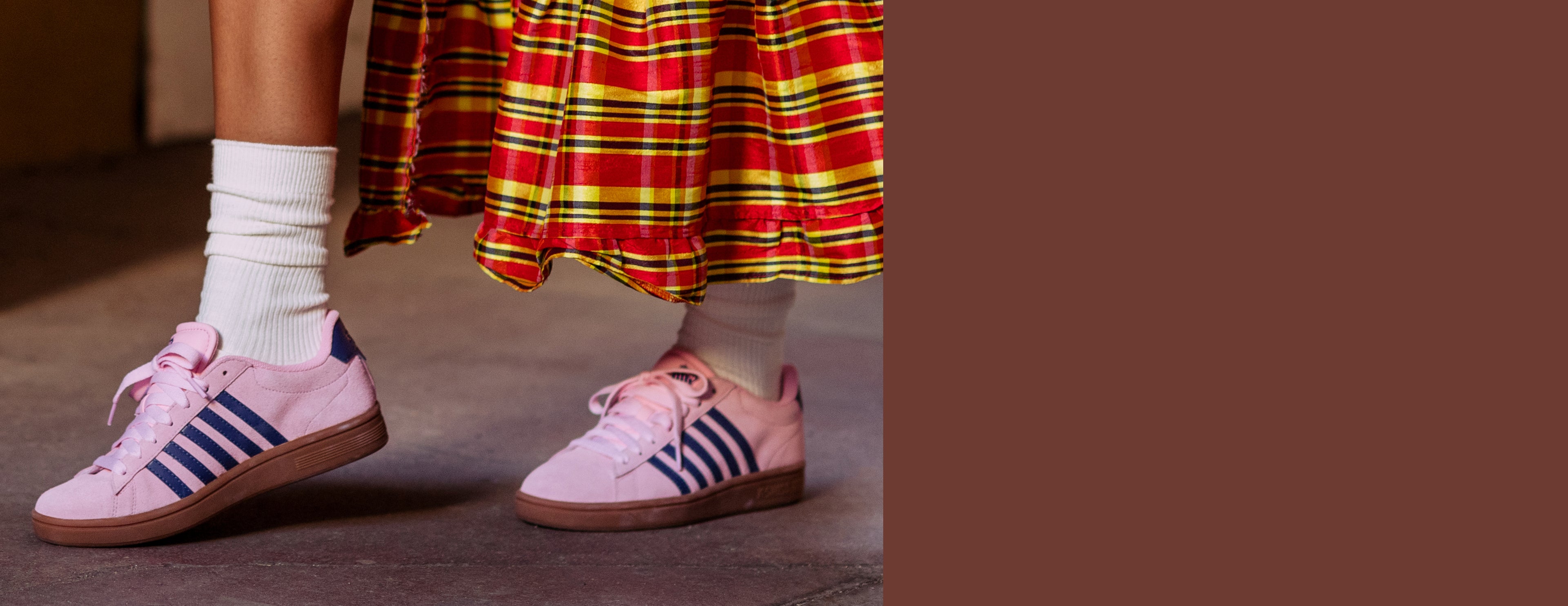 Pink Adidas Originals sneakers with blue stripes, worn with white socks and a red, yellow, and green plaid skirt, shown against a brown background.