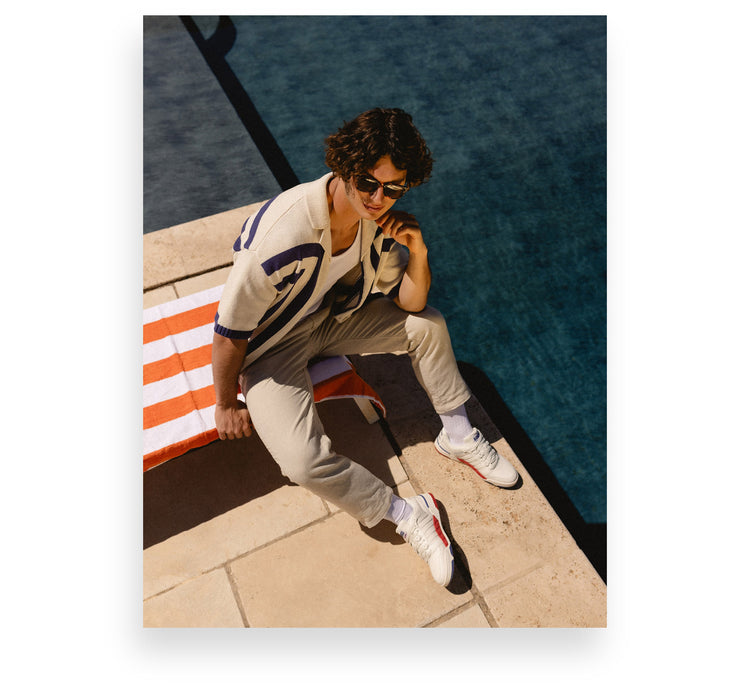 Image of a young man sitting on a lounge chair near a pool wearing preppy clothing and K-Swiss sneakers