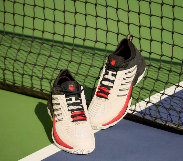 A pair of K-Swiss Hypercourt tennis shoes leaning against a net on a tennis court