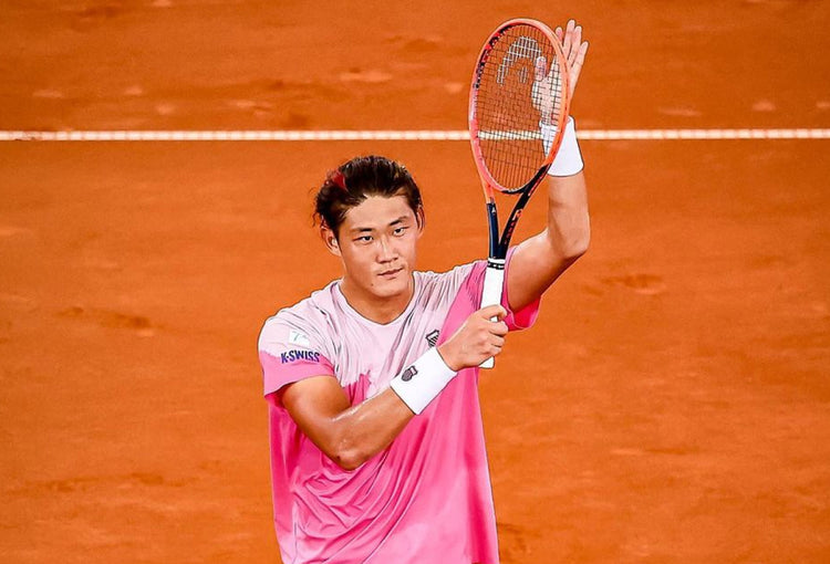 Zhizhen Zhang clapping with a tennis racket on a clay court while wearing a K-Swiss t-shirt and wrist band