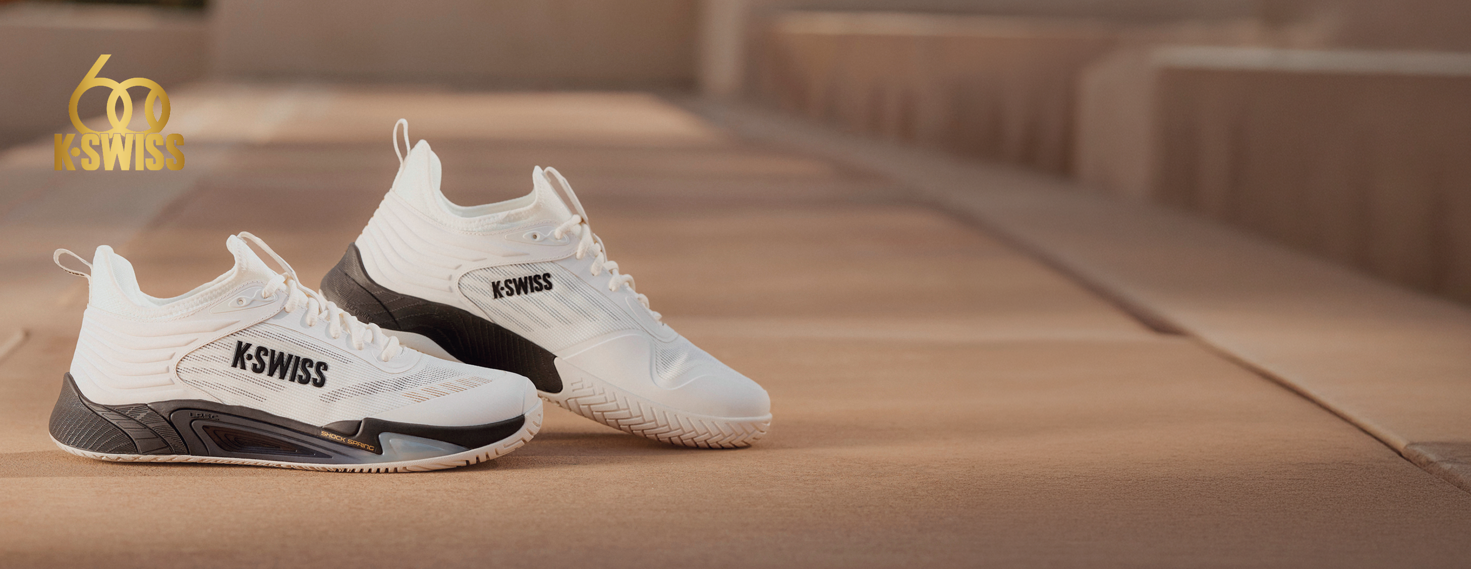 A pair of white K-Swiss athletic sneakers with black and gray accents, displayed on a warm-toned concrete path. The K-Swiss logo is clearly visible on the side of both shoes, and a gold '60 Years K-Swiss' logo is in the top-left corner.