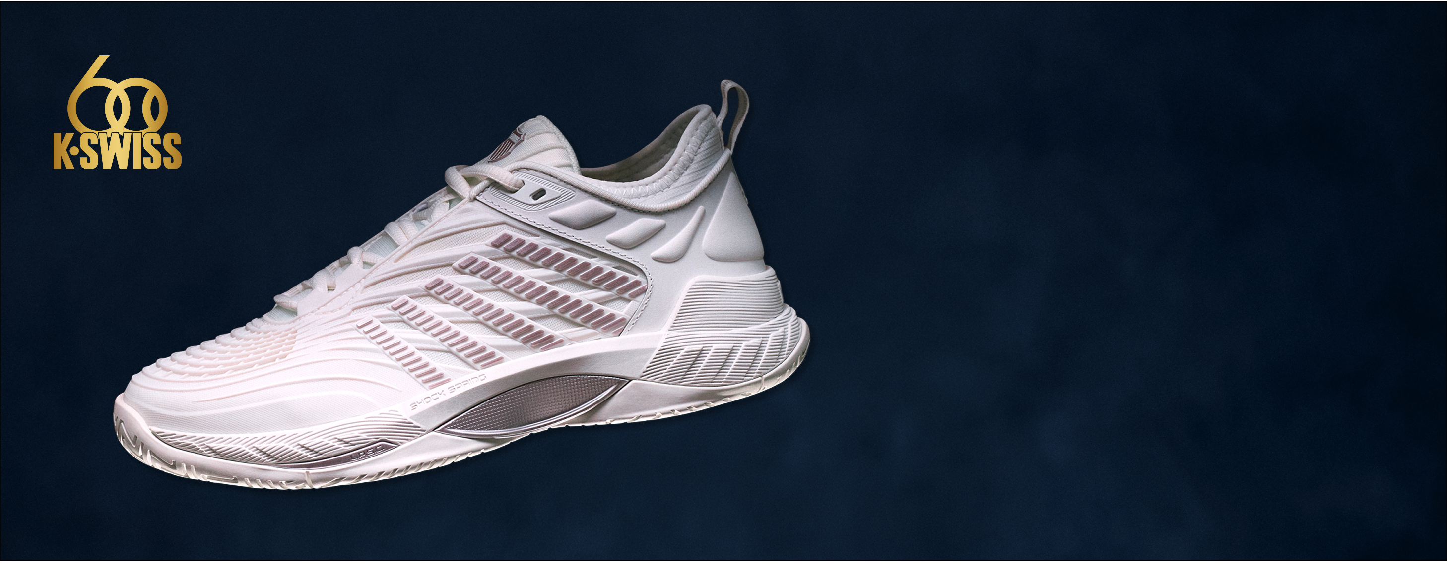A white K-Swiss athletic sneaker with metallic rose-gold patterned side stripes, shown against a dark navy blue background with the K-Swiss logo in the top-left corner.