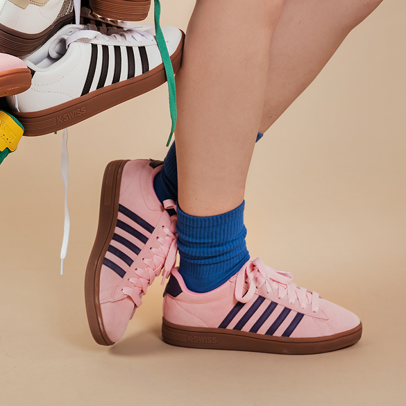 A person wearing pink KSWISS sneakers with dark navy stripes and brown gum soles, paired with blue crew socks, against a warm beige studio background.