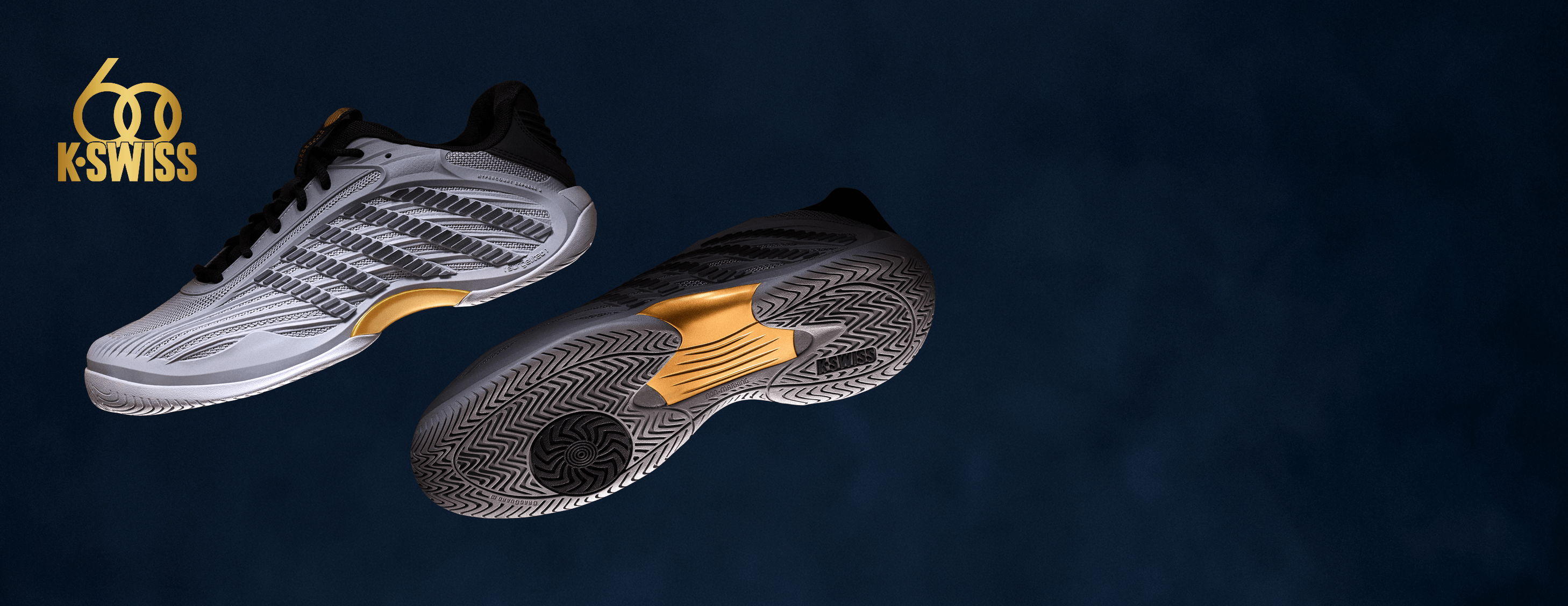 Two K-Swiss indoor soccer cleats shown floating against a dark blue background, one displaying the white and black upper with gold accents, and the other showing the textured black and gray sole.