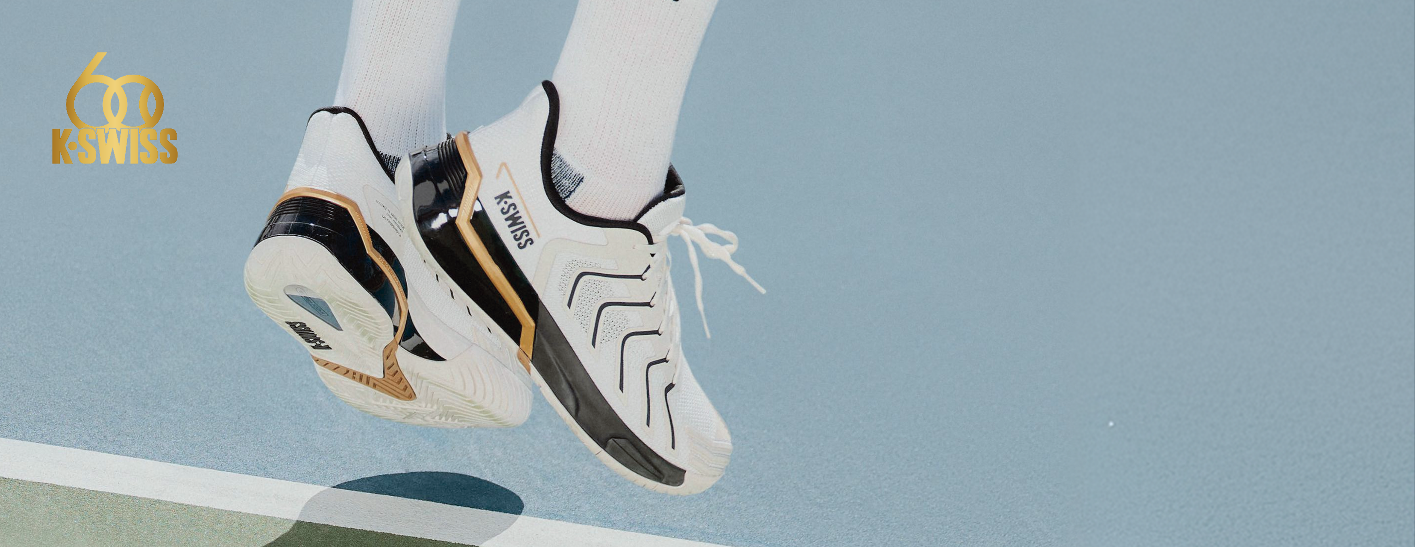 White KINGS athletic sneakers with black and gold accents, worn with white socks on a light blue tennis court.