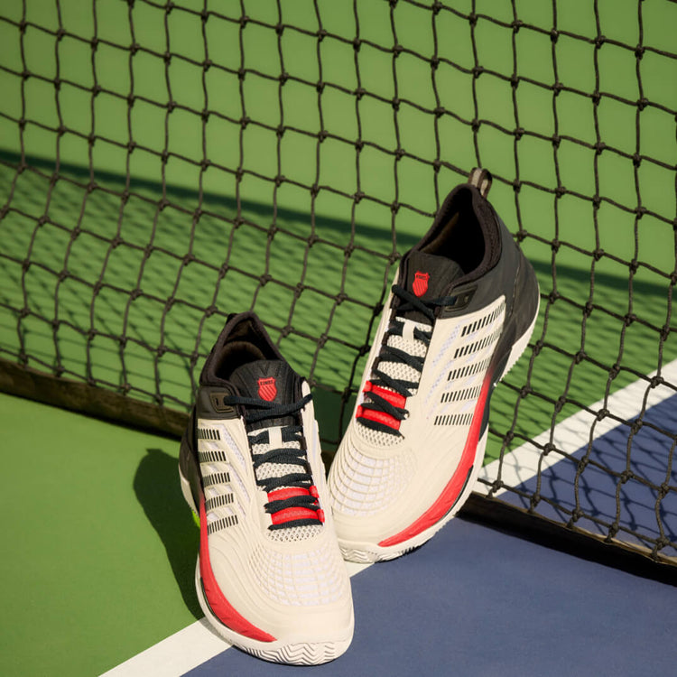 A pair of K-Swiss Hypercourt tennis shoes leaning against a net on a tennis court