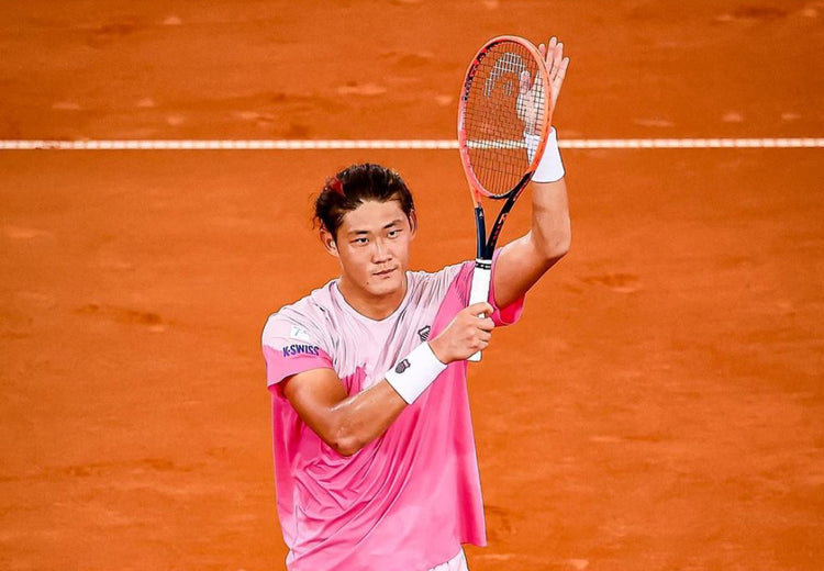 Zhizhen Zhang clapping with a tennis racket on a clay court while wearing a K-Swiss t-shirt and wrist band