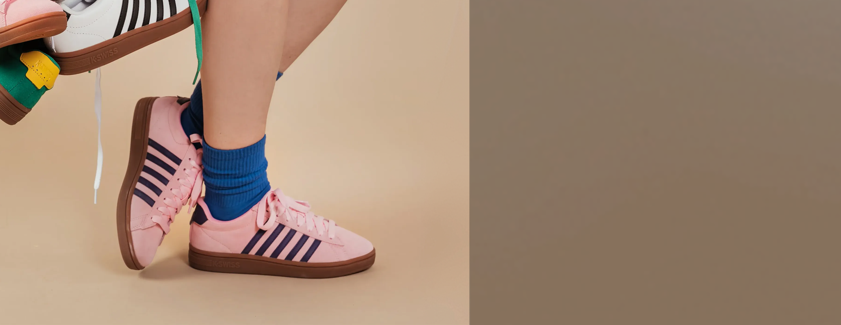 A person wearing pink Kswiss sneakers with dark navy stripes and brown gum soles, paired with blue crew socks, against a warm beige studio background.
